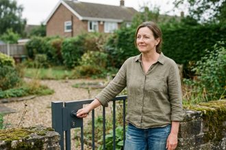 Femme examinant un portail de jardin en acier anthracite dans un jardin suburban avec mur en pierre et végétation naturelle